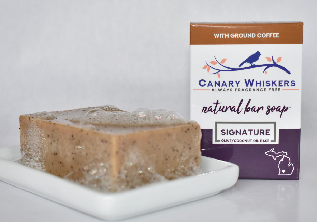 Bar of coffee fragrance-free soap with bubbles on a soap dish next to a boxed coffee soap package.
